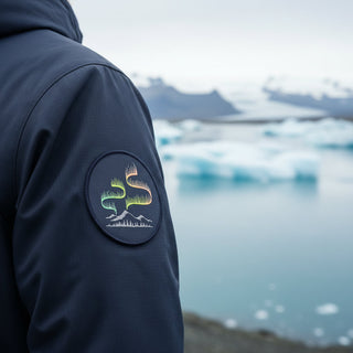Person wearing a dark jacket with a logo patch, standing by a body of water with icebergs, Iceland, patch northern lights 