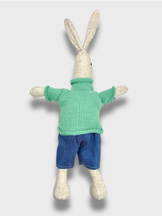 Knitted toy rabbit wearing a green sweater and blue pants on a light gray background