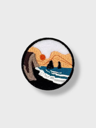 Round iron-on embroidered patch depicting Reynisfjara black sand beach with ocean waves, basalt cliffs, and rock formations in Iceland.
