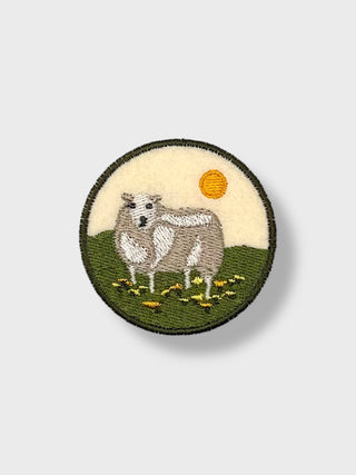 Round iron-on embroidered patch showing an Icelandic sheep standing in a green field under a bright sky.