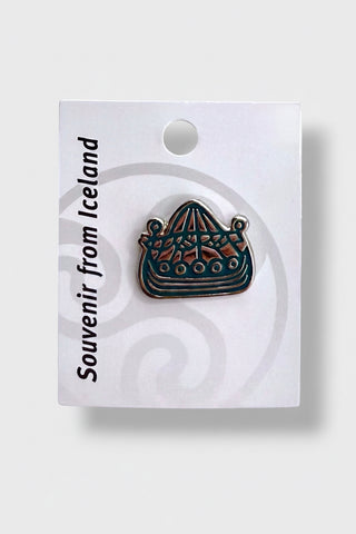 An enamel pin shaped like a sailboat, mounted on a white souvenir card from Iceland. The pin features a dark teal-blue and black base with brown and gold details.