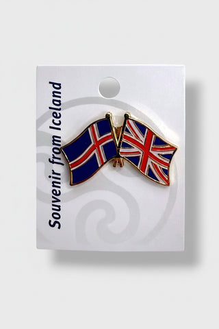 An enamel lapel pin shaped like two small Icelandic flags crossed, attached to a white souvenir card that reads 'Souvenir from Iceland' . The product is photographed on a light gray background.