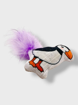 A plush penguin toy with a white and black body, orange beak and feet, and a fluffy purple tail, photographed on a light gray background.