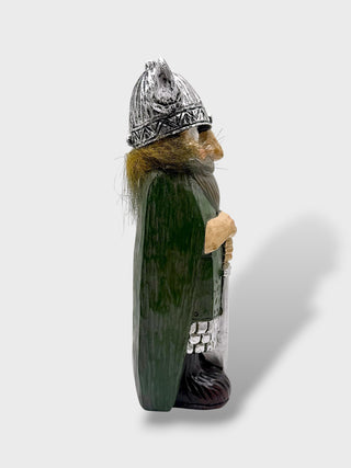 A hand-carved Sami-style dwarf figurine shown in profile against a light gray background. The figurine features a dark green body, a silver-toned metal helmet, brown fur trim, and a silver-toned belt.