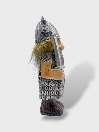A small carved warrior figurine shown in side profile against a light gray background. The figure wears a metallic silver helmet and chainmail armor, has a brown face and beard, and stands on a dark base.