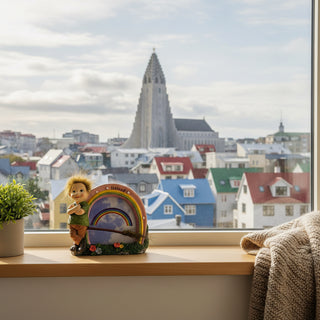 A decorative figurine of a child with wild hair standing beside a rainbow-shaped plaque labeled 'Iceland,' with a broom, grass, flowers, and a cloudy sky backdrop on a light gray background.