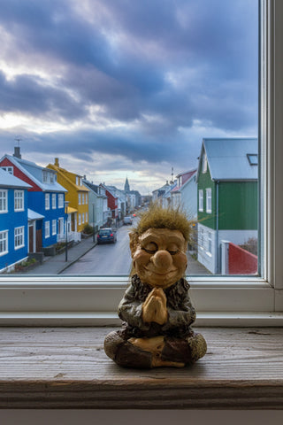 A small figurine of a smiling, meditating gnome or troll with wild brown hair, wearing a dark textured robe and seated cross-legged against a light gray background.