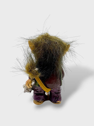 A small fantasy creature figurine shown from the back, featuring wild brown and yellow hair, a dark purple body, and holding a silver-colored sword, set against a plain light gray background.