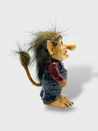A small handcrafted figurine of a fantasy creature with wild dark hair, a long snout, pointed ears, and bare feet, wearing a dark blue outfit and red vest, photographed on a light gray background.