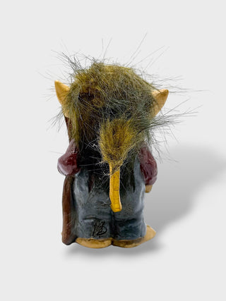 A small fantasy creature figurine shown from the back, with pointed ears, a yellow tail, dark blue-gray body, and shaggy olive-brown hair on a light gray background.