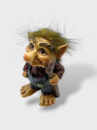 A small fantasy creature figurine with large eyes, pointed ears, wild spiky hair, and bare feet, wearing a dark reddish-brown top and dark pants, photographed on a light gray background.