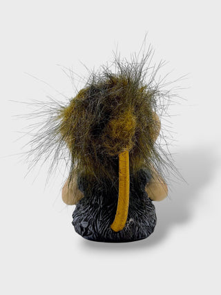 A small sculptural figurine of a wild animal, likely a beaver or similar rodent, shown from the back. It features dark brown and black textured fur, a prominent curved yellow tail, and light tan ears, set against a plain light gray background.