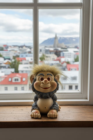 A small seated fantasy creature figurine with a beige face and hands, gray furry body, orange eyes, and spiky tan hair, photographed on a light gray background.