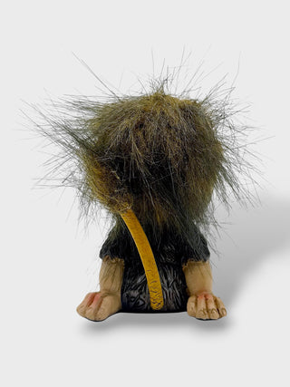 A small figurine or sculpture of a creature with a large, shaggy brown and black mane, pale beige limbs, and a curved yellow tail, shown against a light gray background.
