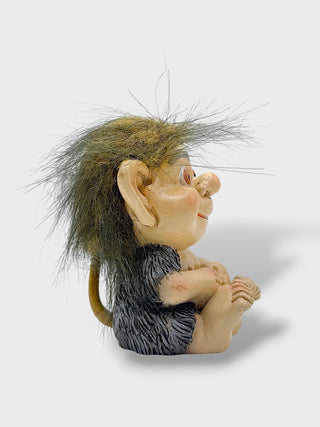 A small sculpted figurine of a seated goblin or troll character with large ears, a long spiky hair, and a dark textured body, photographed on a plain light gray background.