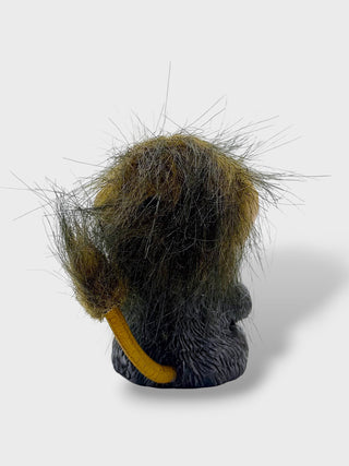 A small sculptural figurine or decorative object shaped like a furry animal head seen from the back, with dark gray and black fur, golden brown highlights, and a curved yellow accent, photographed on a light gray background.