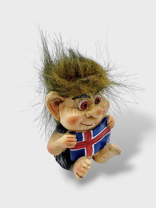 A small figurine of a troll with wild brown hair holding an Icelandic flag, set against a plain light gray background.