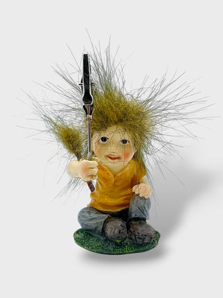 A whimsical gnome figurine with wild grass hair, wearing a yellow shirt and blue pants, holding a pitchfork on a small green base against a light gray background.