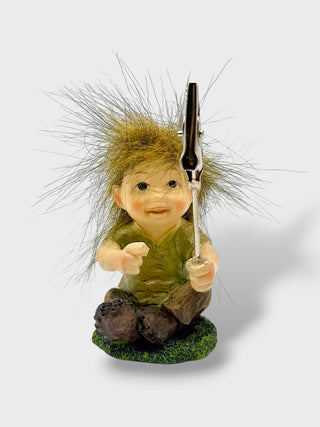 A small gnome figurine with long, spiky brown hair, wearing an olive green shirt and dark pants, sitting on a grassy base and holding a black-and-silver object upright.