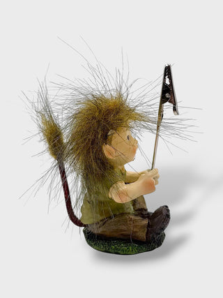 A whimsical figurine of a seated child with long, messy brown hair holding a flagpole with a black flag, standing on a small textured base against a light gray background.