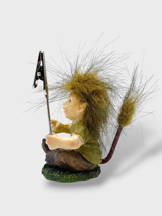 A small decorative figurine of a seated creature with long brown and tan hair, holding a black flag on a golf club, standing on a green textured base against a light gray background.