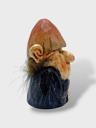 A small carved gnome figurine with a brown hat, beige face and hands, dark blue-black clothing, and brown-and-green facial hair, photographed on a light gray background.