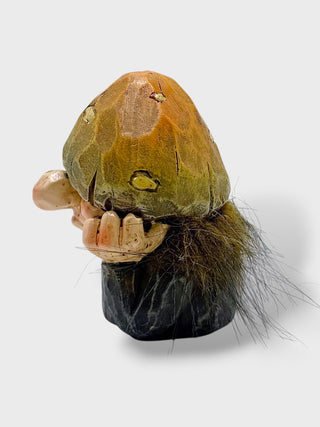 A small sculptural figurine resembling a stylized mushroom or organic form, with a tan and olive-brown top, pale beige protruding side elements, dark brown hair-like strands, and a black base, photographed on a light gray background.