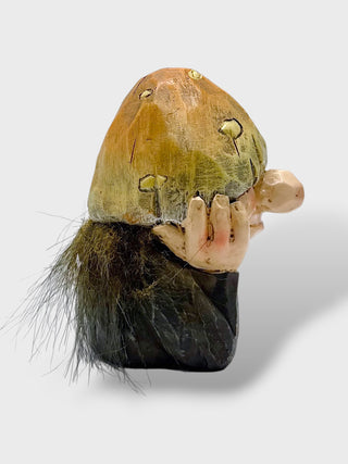 A small sculptural figurine of a mushroom-like cap with pale hands holding it, set against a light gray background.