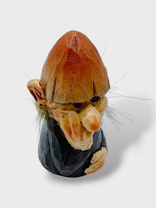 A small sculpted figurine of a gnome or goblin head and upper torso, with a brown conical hat, pale beige face and hands, dark clothing, and short grayish hair, photographed on a light gray background.