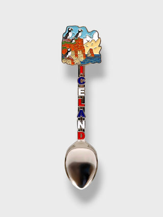A novelty spoon featuring a colorful enamel handle shaped like the map of Iceland with penguins and scenery, and the word 'ICELAND' spelled vertically in red, white, and blue. The spoon bowl is polished silver.