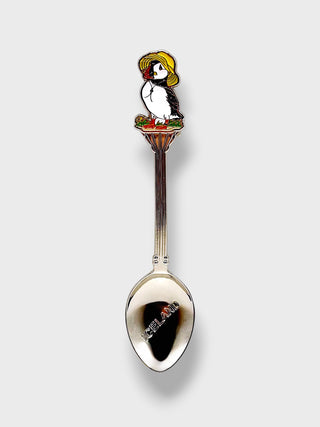 A silver metal spoon with a decorative handle featuring an enamel puffin wearing a yellow hat, set against a light gray background.