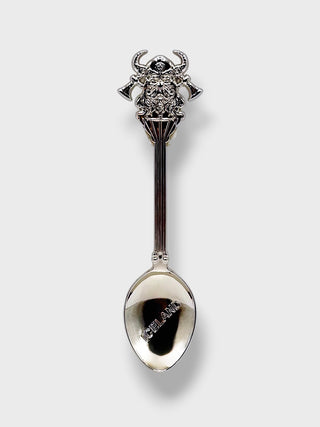 A silver spoon with an ornate sculpted handle featuring a horned animal motif, photographed against a light gray background.