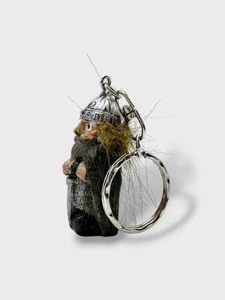 A small decorative keychain featuring a carved dark figure with a silver helmet and beard, attached to a large silver ring with wispy hair-like details, photographed on a light gray background.
