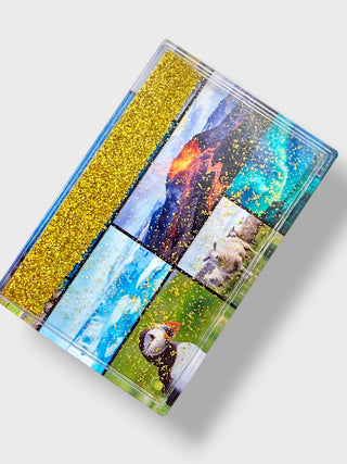 A rectangular clear resin box or tray shown at an angle on a light gray background. The lid features a collage of scenic photographs with gold glitter throughout, including a mountain, water, sheep, and a puffin.