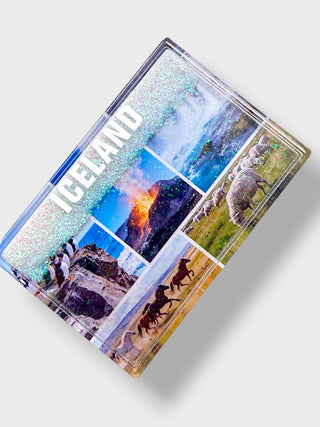A rectangular travel souvenir magnet or plaque featuring a collage of Iceland landscapes and animals, with the word 'ICELAND' printed diagonally in white. The design includes snowy mountains, waterfalls, a volcano, sheep, horses, and glaciers, set against a light gray background.