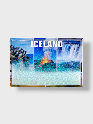 A rectangular souvenir magnet featuring a glittery, stylized depiction of Iceland with a volcano, waterfall, and the word 'ICELAND' in white text across the top, shown on a light gray background.