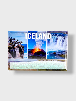 A rectangular souvenir magnet featuring a collage of Iceland landscapes, including penguins on a cliff, an active volcano, and a waterfall, with the word 'ICELAND' in bold white letters. The bottom portion is decorated with a light green and white glittery pattern, and the magnet is shown against a neutral light gray background.