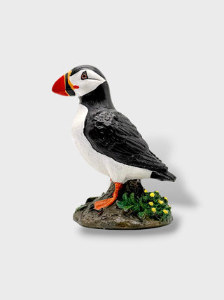 A painted puffin figurine standing on a small rocky base with greenery and yellow flowers, photographed against a plain light gray background.