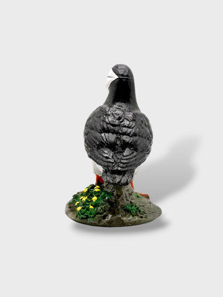 A small decorative bird figurine standing on a textured base with greenery and yellow flowers, photographed against a plain light gray background.