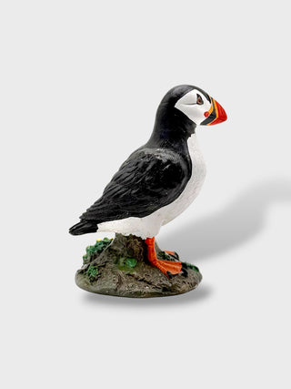 A small puffin figurine standing on a textured base, photographed against a plain light gray background. The bird has black and white coloring with bright orange feet/beak and a small red accent.