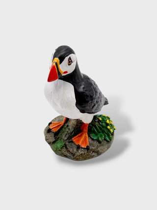 A painted puffin figurine standing on a rocky base with green seaweed and small yellow flowers, photographed against a plain light gray background.