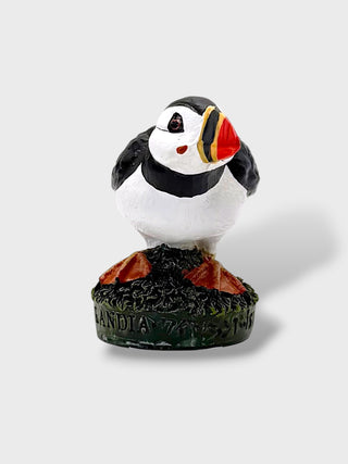 A small decorative puffin figurine standing on a dark textured base with the word 'LANDIA' visible on the front, photographed against a light gray studio background.