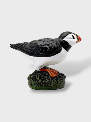 A decorative puffin figurine standing on a textured green base with the words 'ISLANDS ARE MY BUSINESS' embossed on the front. The bird has a black and white body with a bright orange beak and feet, photographed against a clean light gray background.