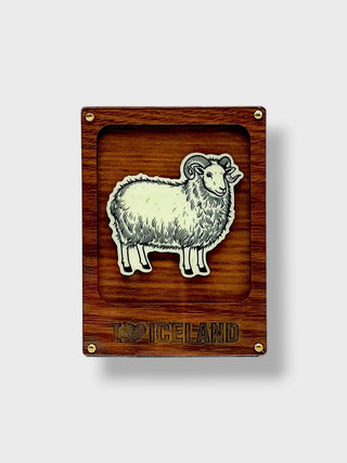 A rectangular wooden plaque featuring a raised white sheep illustration in the center and the word 'ICELAND' engraved at the bottom, with small brass corner screws, shown on a light gray background.