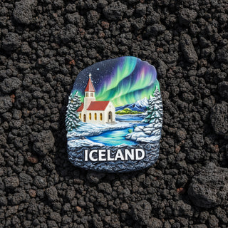 An irregularly shaped souvenir magnet or plaque featuring a winter landscape of Iceland with a church, snowy trees, mountains, and a vivid aurora borealis sky. The word "ICELAND" is printed in white at the bottom.