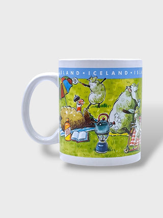 A white ceramic mug featuring a colorful cartoon illustration of sheep and picnic items on a grassy field, with the text 'ICELAND' printed in blue around the top band.