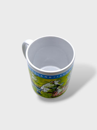 A white ceramic mug with a colorful illustration of Iceland landscapes and text, photographed from an overhead angle on a light gray background.