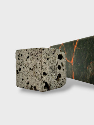 A close-up of a textured gray stone block with dark volcanic pores in the foreground, with a dark green marble slab showing orange veining in the background, set against a light gray studio backdrop.
