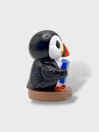 A stylized puffin figurine sitting on a round brown base, holding a blue object. The puffin has a black body, white face, orange beak, and orange feet, photographed against a light gray background.