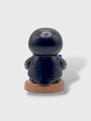 A black penguin figurine sitting on a round brown base, photographed from the back against a light gray background.